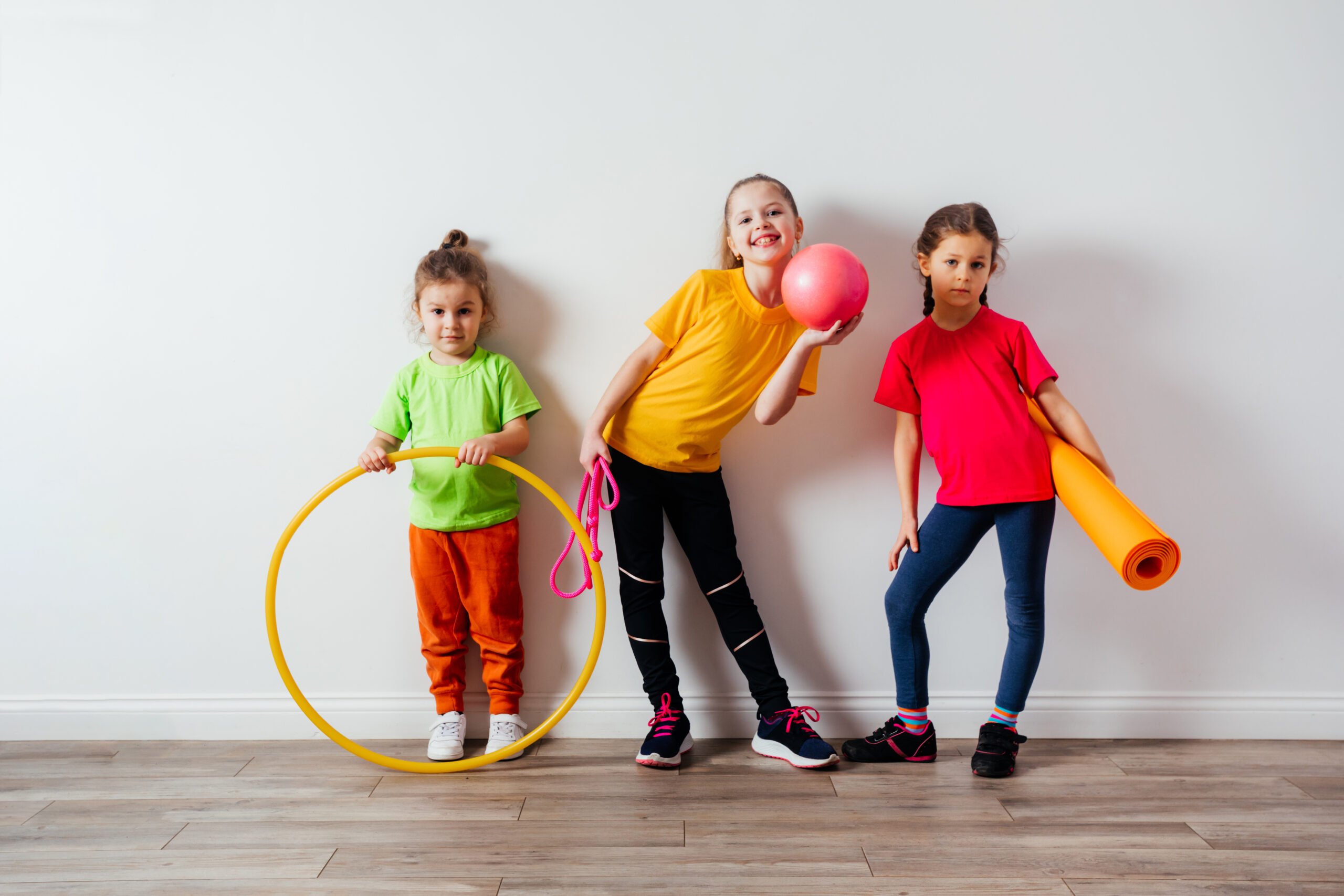 Two pretty sportive girls and little boy posing with various sport equipment on white wall background. Active children in colorful t-shirts with ball, dumbbells, skipping rope and hula hoop.